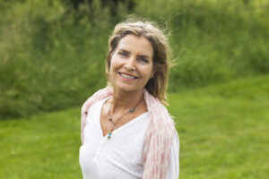 kvinne smiler mot kamera, hun har på hvit genser, grønt gress i bakgrunnen