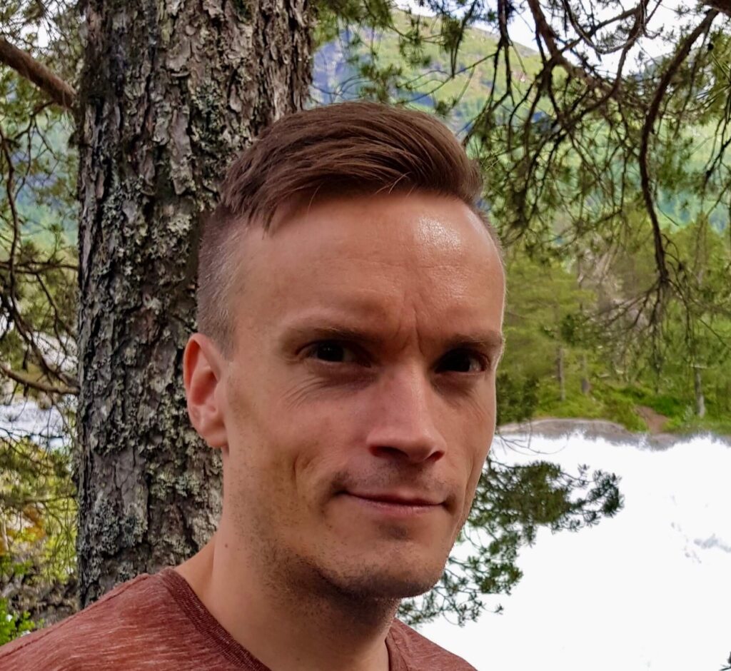 portrett av mann med kort hår, trær i bakgrunnen, smiler litt