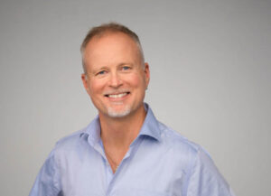 middelaldrende mann smiler mot kamera, grå bakgrunn, han har lys skjorte på